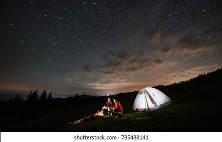 Night camping in the mountains. Couple tourists have a rest at a campfire near illuminated tent under amazing night starry sky. Low light