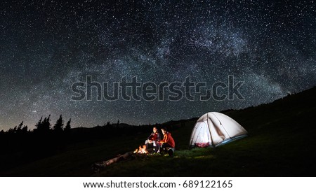 Night camping in the mountains. Couple tourists have a rest at a campfire near illuminated tent under amazing night sky full of stars and milky way. Low light. Picture aspect ratio 16:9