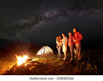 Night camping in mountains. Tourists standing in front of burning bonfire. Illuminated tent and starry sky on background. Woman holding in arms small daughter. Tourism and recreation concept. - Powered by Shutterstock