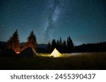 Night camping in mountains under starry sky. Tourist tents in campsite near burning campfire under beautiful sky full of stars with Milky way above forest. Concept of tourism and traveling.