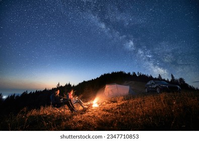 Night camping in the mountains under starry sky with Milky way. Travelling couple having a rest at campfire, tourist tent and off-road vehicle in the evening on mountain lawn and stargazing together.