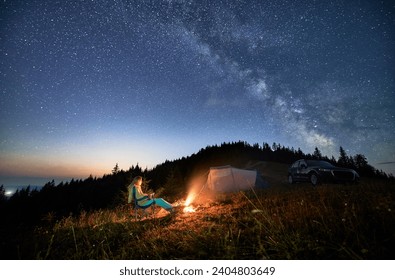 Night camping in the mountains under starry sky full of stars. Travelling woman admiring starry night in mountains. Young woman sitting next to camp fire, tourist tent and off-road car. - Powered by Shutterstock