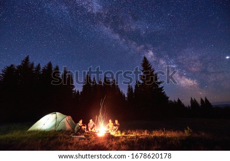 Night camping near bright fire in spruce forest under starry magical sky with milky way. Group of four friends sitting together around campfire, enjoying fresh air near tent. Tourism, camping concept.