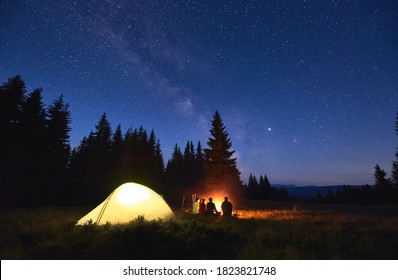 Night Camping Near Fire, Forest And Mountains On Background. Group Of Friends Warming Up Near Bright Bonfire. People Sitting Near Tourist Illuminated Tent Under Night Sky Full Of Stars And Milky Way.