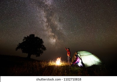 Night Camping Near Silhouette Of Big Tree On Grassy Valley. Tourist Man Standing Near Burning Campfire, Pointing At Beautiful Starry Sky And Milky Way To Young Woman Sitting In Glowing Tent Entrance