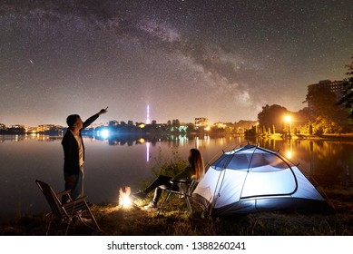 Night camping on lake shore. Couple tourists having a rest near tent and campfire. Man pointing to beautiful night sky full of stars and Milky way, city lights on background. Outdoor lifestyle concept