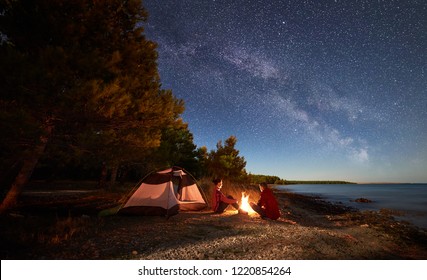 Night camping on shore. Man and woman hikers having a rest in front of tent at campfire under evening sky full of stars and Milky way on blue water and forest background. Outdoor lifestyle concept