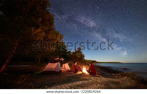 Night camping on shore. Man and woman hikers\
having a rest in front of tent at campfire under evening sky full\
of stars and Milky way on blue water and forest background. Outdoor\
lifestyle concept