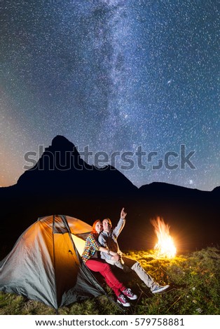Night camping. Pair of hikers sitting near tent and campfire and enjoying incredibly beautiful starry sky in the background silhouette of the mountains. Long exposure