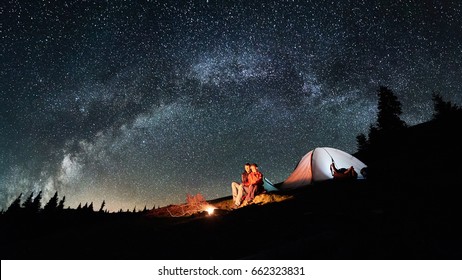 Night camping. Romantic couple tourists have a rest at a campfire near illuminated tent under amazing night sky full of stars and milky way. Astrophotography. Picture aspect ratio 16:9