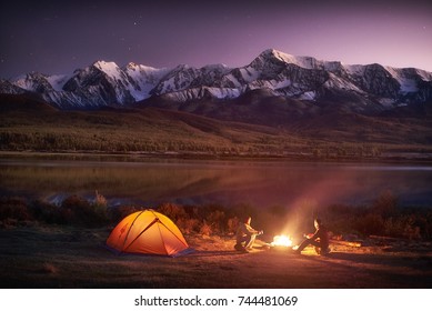 Night camping. Two men tourists sitting at the illuminated tent near campfire under amazing sunset evening sky in a mountains area. Snow mountain in the background