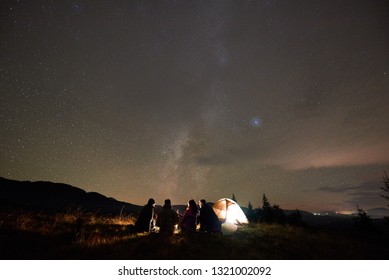 Night Camping Under Stars, Tourism And Active Lifestyle. Back View Silhouette Of Four Persons Sitting On Grassy Valley At Tourist Tent And Burning Campfire On Copy Space Background Of Dark Starry Sky.