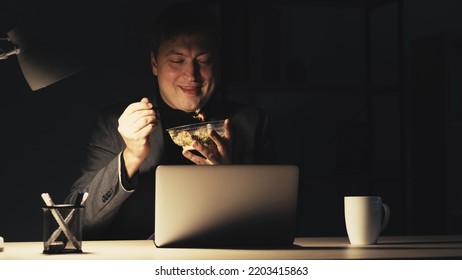 Night Dinner. Happy Man. Enjoying Meal. Funny Hungry Business Guy Sitting Desk With Laptop Eating Salad In Dark Room Interior.