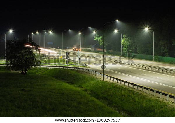 Night expressway in the light of\
streetlights. A truck is parked on the side of the road. There are\
trees and green grass at the edges of the road. Night\
scenes