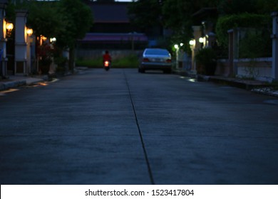 Night Fall In A Gated Community. Quiet Street In A Village. Concrete Road With Lights From House Wall Lamp. Blurred Car Parking Along The Lane. Urbanisation Sunset Scene.