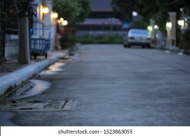 Night Fall In A Gated Community. Quiet Street In A Village. Concrete Road With Lights From House Wall Lamp. Blurred Car Parking Along The Lane. Urbanization Sunset Scene.