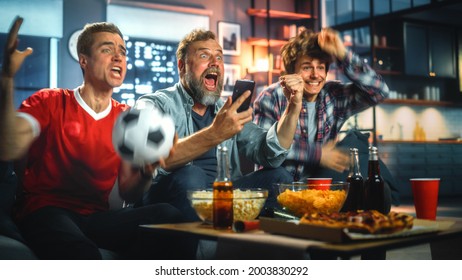 Night At Home: Three Soccer Fans Sitting On A Couch Watch Game On TV, Use Smartphone App To Online Bet, Celebrate Victory When Sports Team Wins. Friends Cheer Eat Snacks, Watch Football Play.