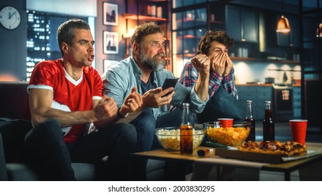 Night At Home: Three Soccer Fans Sitting On A Couch Watch Game On TV, Use Smartphone App To Online Bet, Celebrate Victory When Sports Team Wins. Friends Cheer Eat Snacks, Watch Football Play.