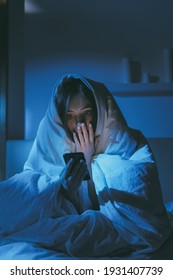 Night insomnia. Late online. Stress anxiety. Social media addiction. Terrified scared girl reading disturbing Internet news on mobile phone sitting on bed in blanket in dark bedroom with blue light.