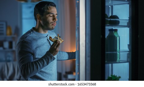 At Night In The Kitchen Handsome Attractive Young Man Is Eating A Leftover Pizza From The Fridge. He Is Hungry And Feels Satisfied.