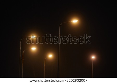 Night lampposts shines with faint mysterious yellow light through evening fog. Streetlights shine at quiet city night, magic atmospheric light in mystical darkness