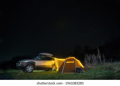 Night Landscape With Illuminated Tent And 4wd Car, Lights And Stars In The Background. Spring Camping. Tourism And Vacation. Roof Rack, Car Luggage Box