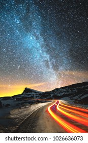 Night Landscape. Night Sky With A North Hemisphere Milky Way And Stars. The Night Road Illuminated By The Car Winds With A Serpentine And Leaves In A Distance To A Foot Of An Acute Rock. Light Trails