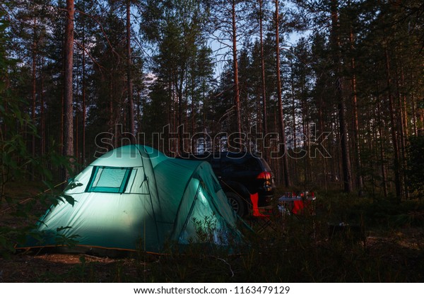 Night
landscape with a tent in the forest near lake. The light from the
lantern in a tent. Car and portable table and chairs, green tourist
tent. Romantic evening with a tent at
night.