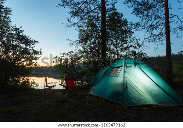 Night\
landscape with a tent in the forest near lake. The light from the\
lantern in a tent. Car and portable table and chairs, green tourist\
tent. Romantic evening with a tent at\
night.