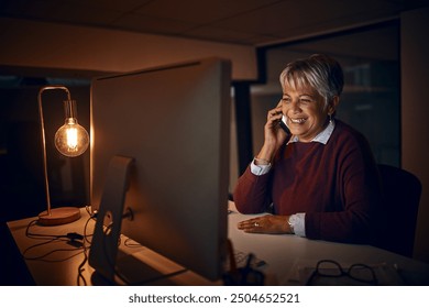 Night, mature woman and phone call at computer for business, talk o rcontact in office. Dark, happy or lady at desk with tech, scheduling or chatting on mobile for funny story or gossip on late shift - Powered by Shutterstock