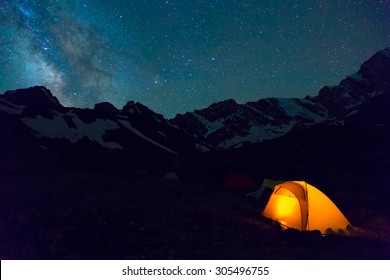 Night Mountain Landscape With Illuminated Tent. Silhouettes Of Snowy Mountain Peaks And Edges Night Sky With Many Stars And Milky Way On Background Illuminated Orange Tent On Foreground