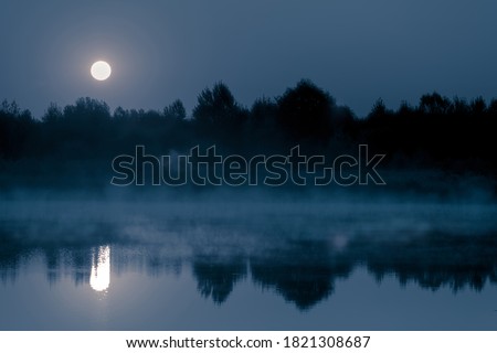 Night mystical scenery. Full moon over the foggy river and its reflection in the still water.