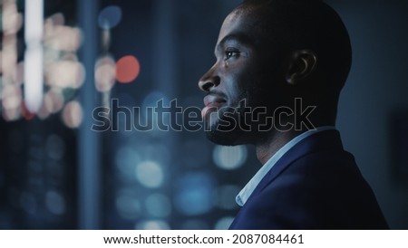 Night Office: Stylish Close-up Portrait of Powerful Black Businessman Wearing Suit Standing, Looking out of the Window on a Big City. Ambitious African CEO Thinking of e-Commerce Investment