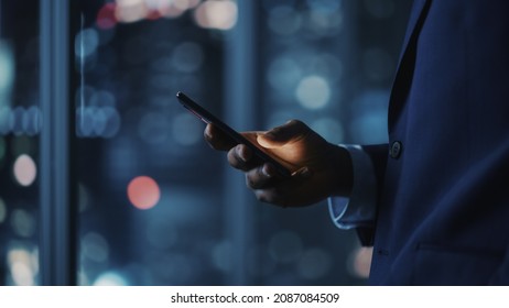 Night Office: Successful Black Businessman Wearing Suit Standing, Using Smartphone. CEO Browsing Internet, Managing Social Media Strategy in e-Commerce Software. Focus on Hand with Mobile Phone