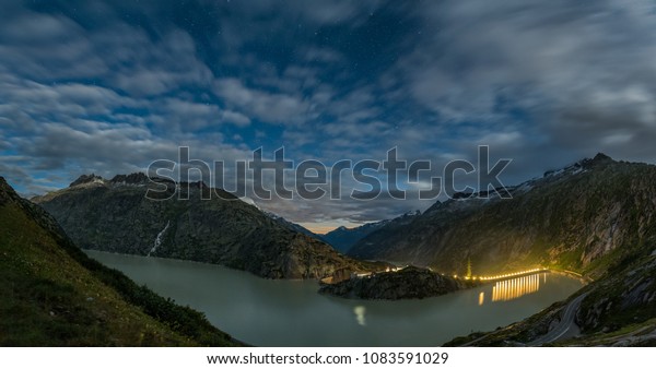 Night panorama  with long exposure of blurred\
moving clouds and still stars on night sky taken in Swiss Alps on\
Grimsel lake.