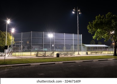 Night Photo Of An Empty City Park Baseball Field At Night With The Lights On As Seen From The Parking Lot And The Bleachers Behind Home Plate.