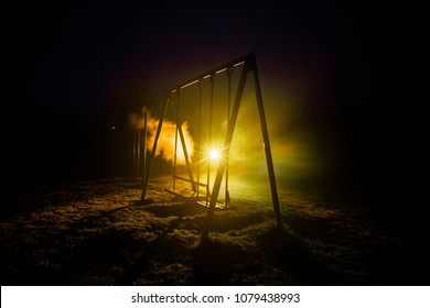 Night photo of metal swing standing outdoor at night time with fog and surreal toned light on background. Nobody there. Lonelyness concept