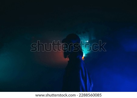 Night portrait of an attractive woman in casual clothes and a cap standing on a foggy street at night in colored light and looking at the camera with a serious face.