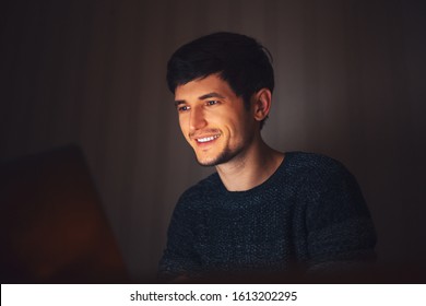 Night Portrait Of Young Happy Man Looking In Laptop In Dark Room At Home.