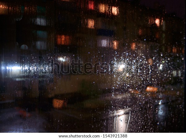 Night rainy sityscape outside the window pane\
- defocused blurred background with water drops on glass, bokeh of\
night illumination, parked cars. Melancholy or romantic mood\
concept, meteorology