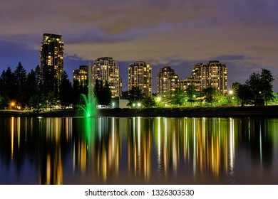 Night scape city view, Coquitlam, Greater Vancouver area, Canada. Fountain in lake and water reflections of modern buildings windows lights - Shutterstock ID 1326303530
