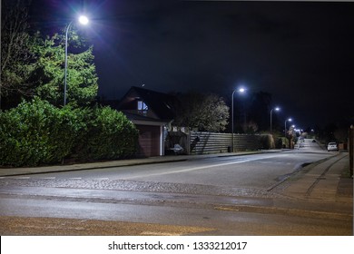 A night scape with an empty residential street lit by pale lamps with small houses on the sides with green bushes