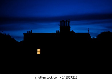 Night Scene Of Only One House Window With Light Swithced On. Chimneys Against Blue Sky 