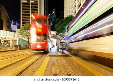 Night Scenery Of Downtown Hong Kong With A Double-decker Tram Moving Fast On The Tracks Amid Highrise Office Towers & A Bus Traveling Under A Pedestrian Footbridge Near The Landmark Square In Hongkong