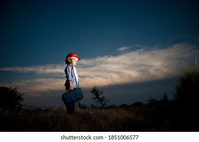 Night Shift Of Little Boy In Red Helmet With Tool Box Standing In Dusk Field With Clouds Lit With Light Of Factory