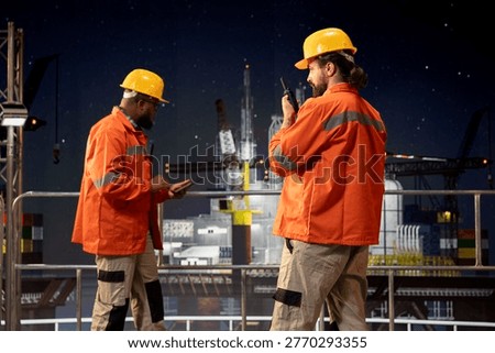 Night shift offshore rig worker using walkie talkie to coordinate safety protocols with logistics crew. Drilling barge professional speaking into handheld radio device while supervising deck machinery