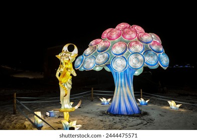 Night show of light installations with light sculptures of tree and a magical creature holding a cat surrounded by smaller sculptures in the shape of flowers against a winter dark background