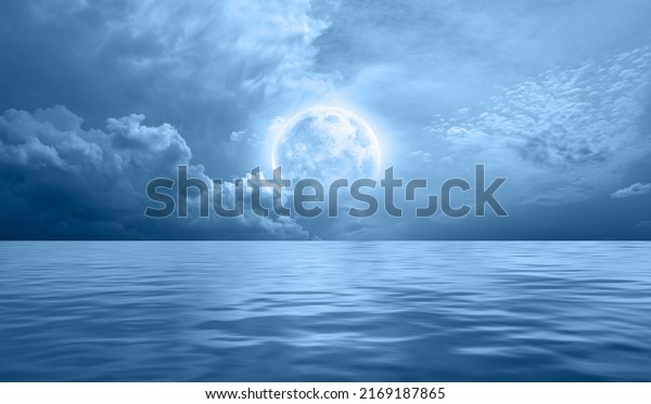 Night sky with blue moon in the clouds over the calm
blue sea, many stars in the background  