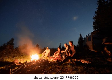 Night sky over mountain hill with car and male hikers near campfire. Group of travelers sitting near bonfire under beautiful blue sky with stars. Concept of night camping, hiking and travelling. - Powered by Shutterstock