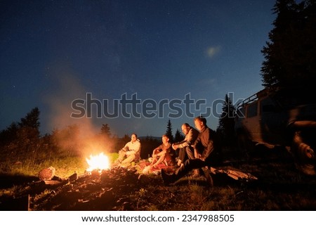 Night sky over mountain hill with car and male hikers near campfire. Group of travelers sitting near bonfire under beautiful blue sky with stars. Concept of night camping, hiking and travelling.